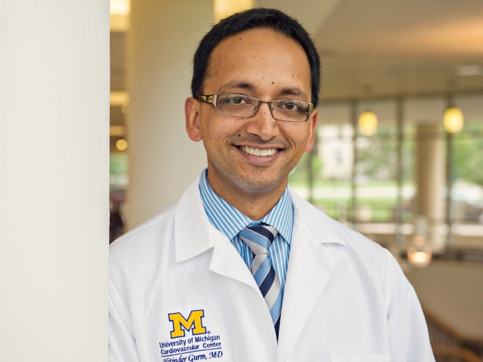 Hitinder Gurm, M.D. wearing a blue shirt, striped tie, and a Michigan Medicine branded white lab coat, standing next to a pillar