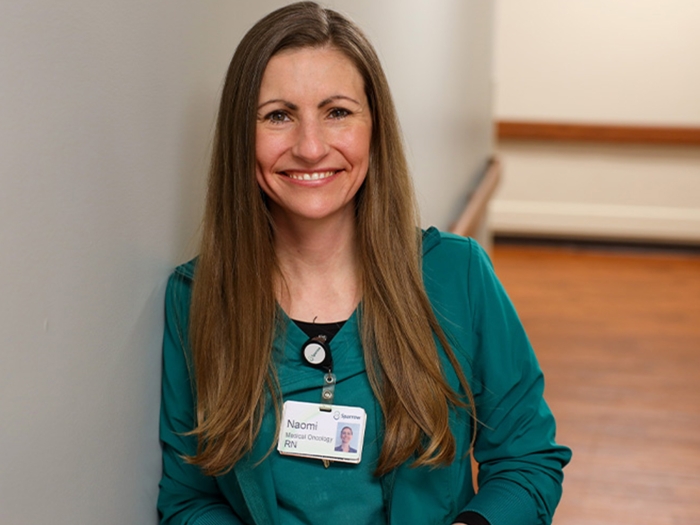 woman with brown long hair and green top and ID badge on smiling at camera against gray wall with brown floor behind and brown thin banisters on wall 