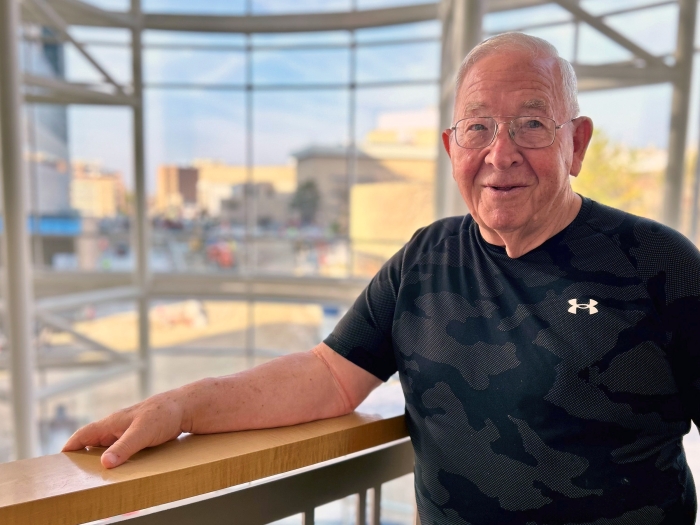 older man with glasses standing at balcony with back to glass windows 