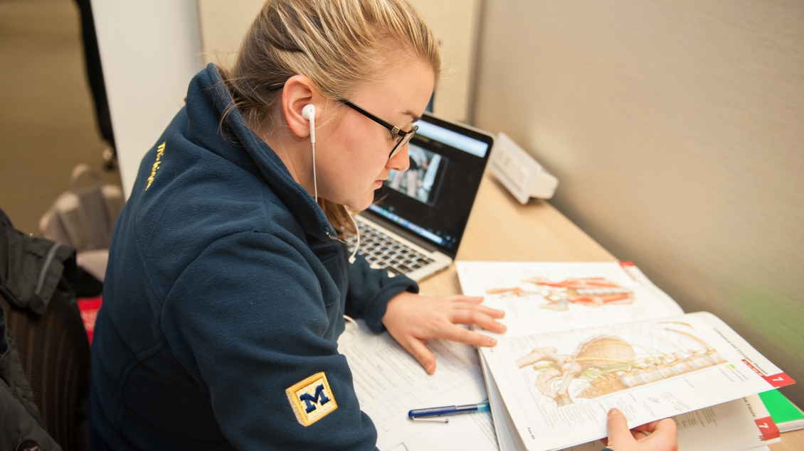 Female student wearing earbuds and Michigan hoodie looking at anatomy textbook with laptop in background