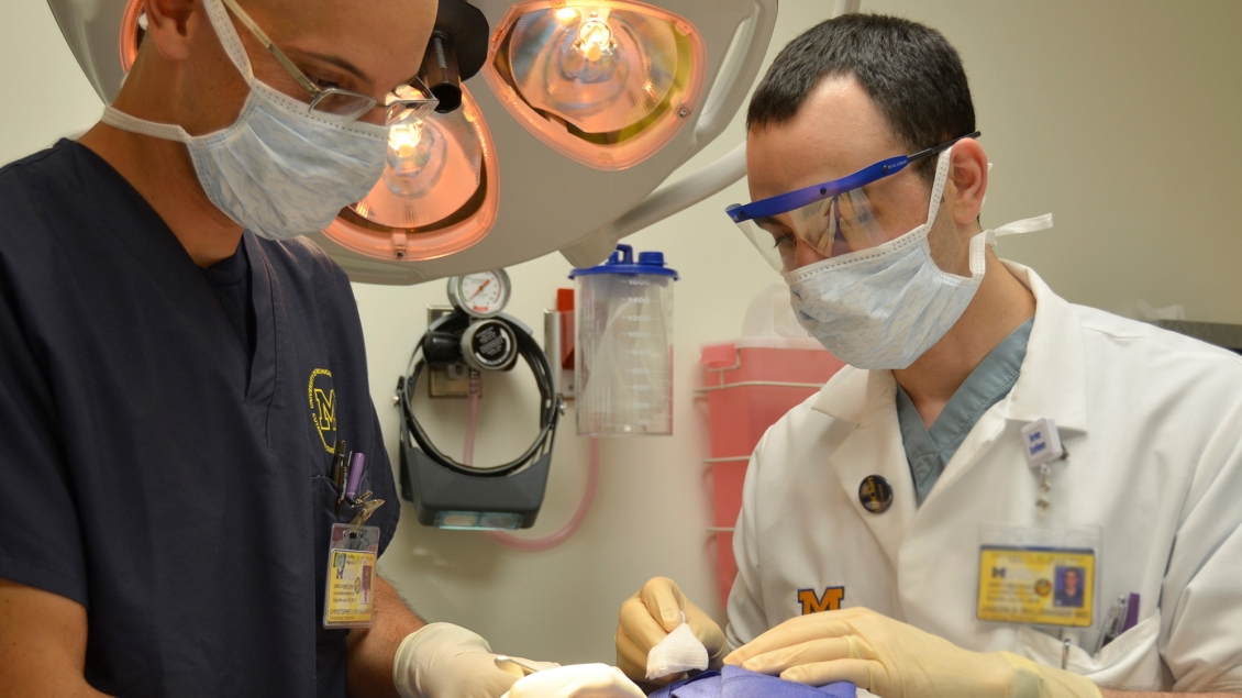 Two providers in masks and gloves perform a procedure on a patient in a dental clinic, with overhead surgical lights illuminated