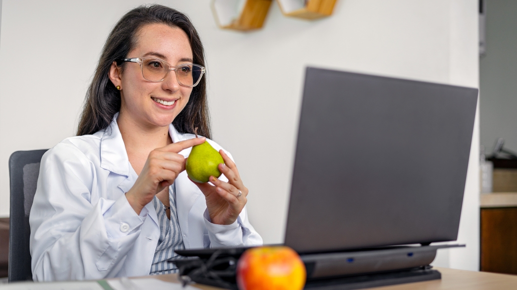 person in white coat at laptop green fruit in hand