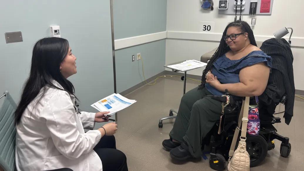 A medical student with a standardized patient who uses a wheelchair have a conversation in an exam room