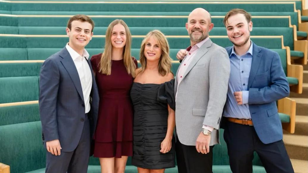 Smiling family photo of 5 members of the Kripke family. They appear to be dressed for a semi-formal event and are standing in an open space with green-cushioned seating visible behind them.