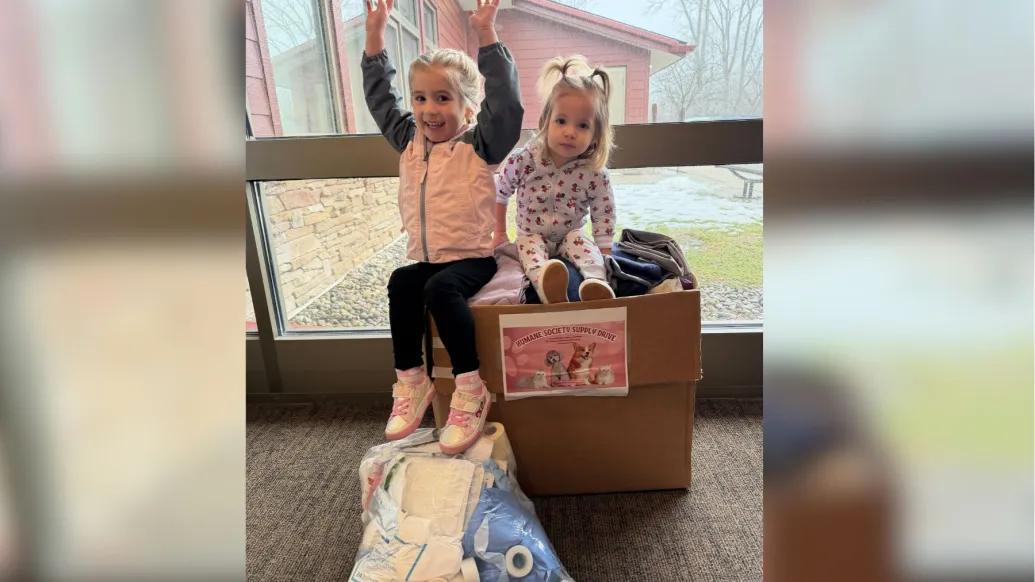 Two little girls sit on a box of donated blankets and towels for a humane society.