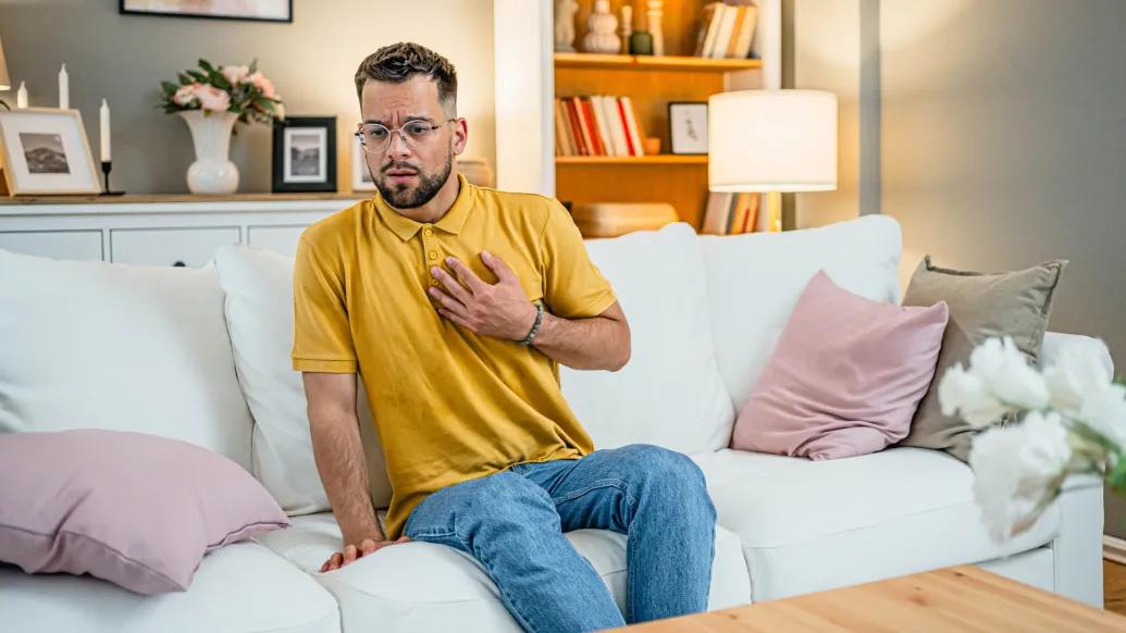 man holding chest with yellow shirt sitting on white couch concerned