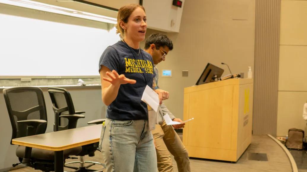 Two people stand at the front of a lecture hall, one wearing a Michigan Medical School t-shirt and gesturing, near a podium with the University of Michigan logo.