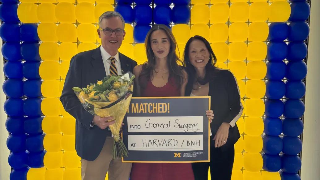 Alt text: **Three people pose in front of a maize-and-blue balloon wall; the person on the left holds a bouquet, and they hold a sign reading “MATCHED! into General Surgery at HARVARD / BWH” with University of Michigan Medical School branding.
