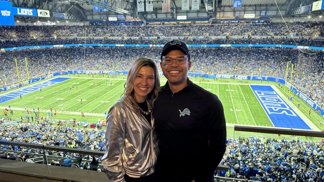 Two people pose in the foreground at an indoor Detroit Lions football game, with a packed stadium and the field visible behind them; one wears a shiny silver jacket and the other a black Lions pullover and cap.