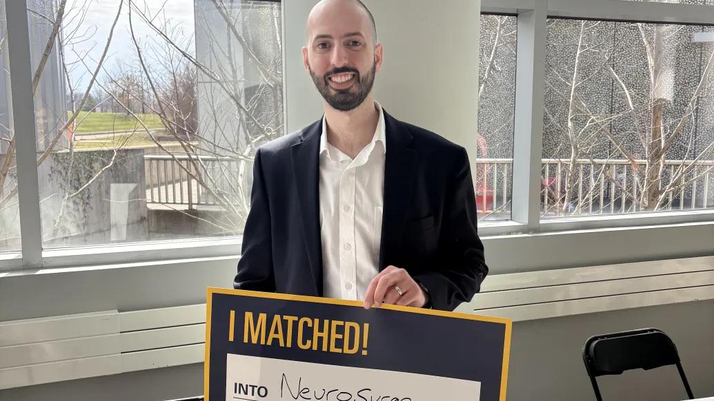Person in a dark blazer and white shirt holds a sign reading “I MATCHED!” with “Into Neurosurgery” handwritten, standing indoors by large windows.