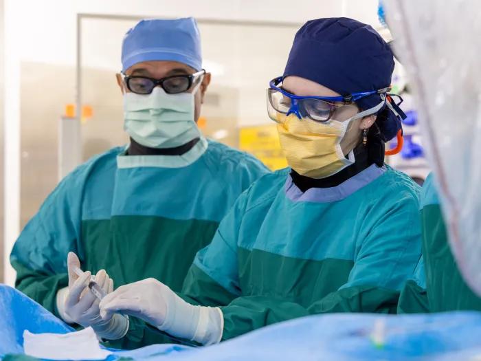 Two gowned and masked doctors wearing glasses, one holding a syringe 