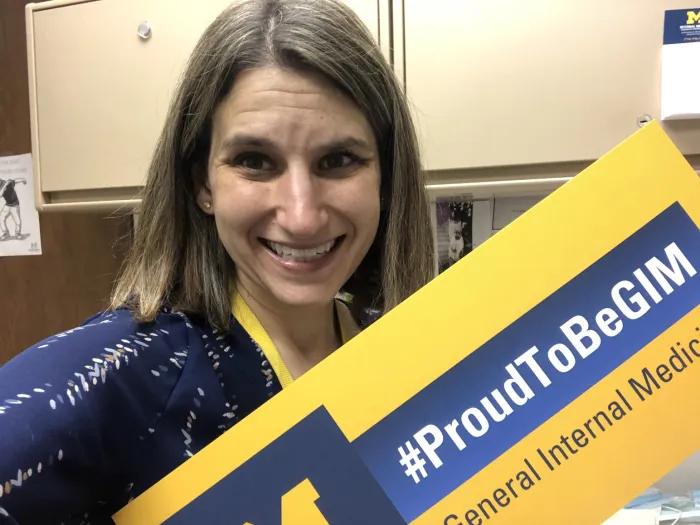 A woman, Alli Ruff, holds a maize and blue sign that reads #ProudToBeGIM, General Internal Medicine