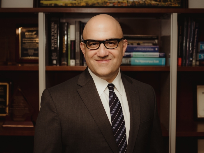 George Mashour, M.D., Ph.D. standing in front of a bookcase, smiling. He is wearing a dark suit, white shirt, dark striped tie and dark framed glasses.