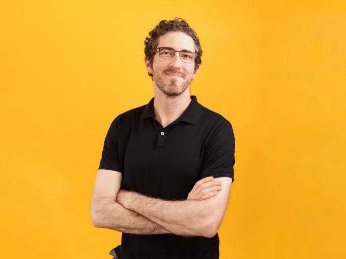 man in black polo folded arms smiling no teeth bright orange background