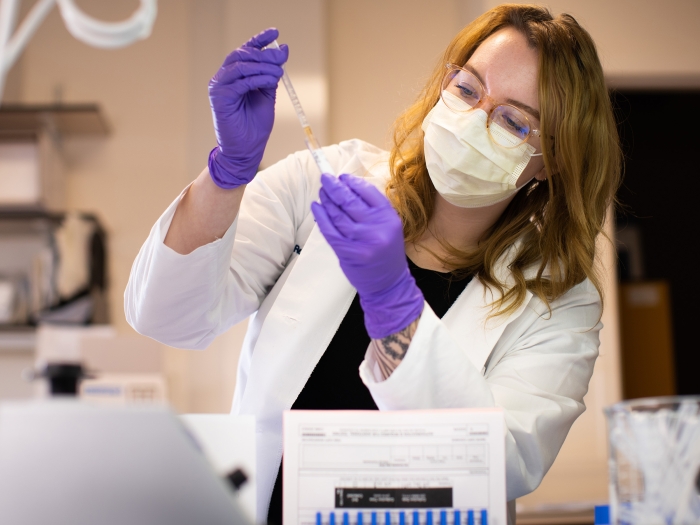 woman putting something in vial in lab coat and purple gloves, glasses and mask in clinical looking area