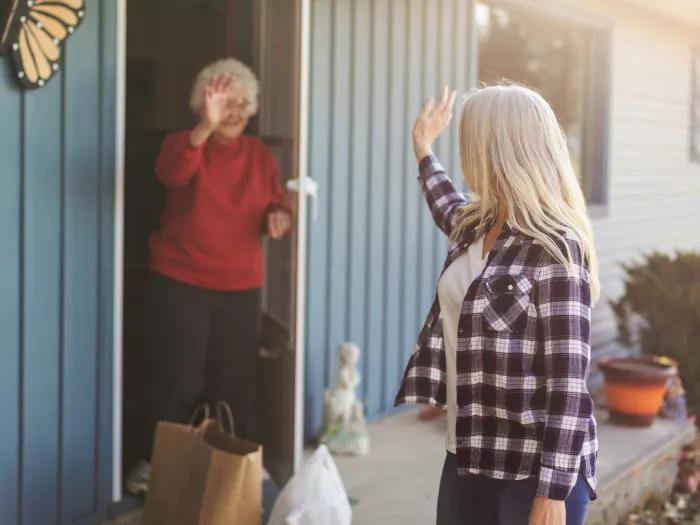 older woman in red long sleeve shirt waving from open door of blue house with other woman leaving with long silver hair and checkered shirt with white undershirt turned towards the door