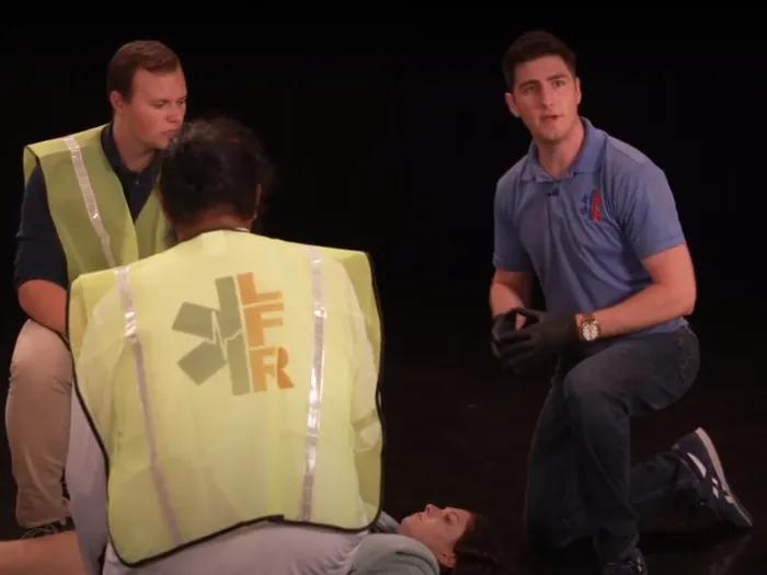Man in a blue polo shirt kneeling on a stage, with two men in neon yellow safety vests kneel next to him, over another person lying on the floor