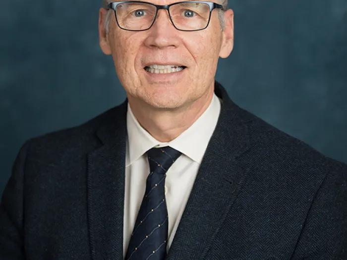 Man with grey hair and glasses wearing a black blazer and tie.
