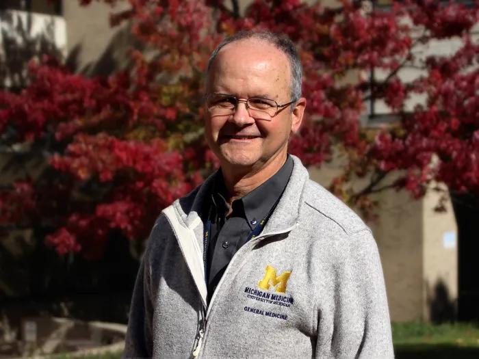 Man wearing glass and a grey Michigan zip-up sweater standing in front of red-leafed trees