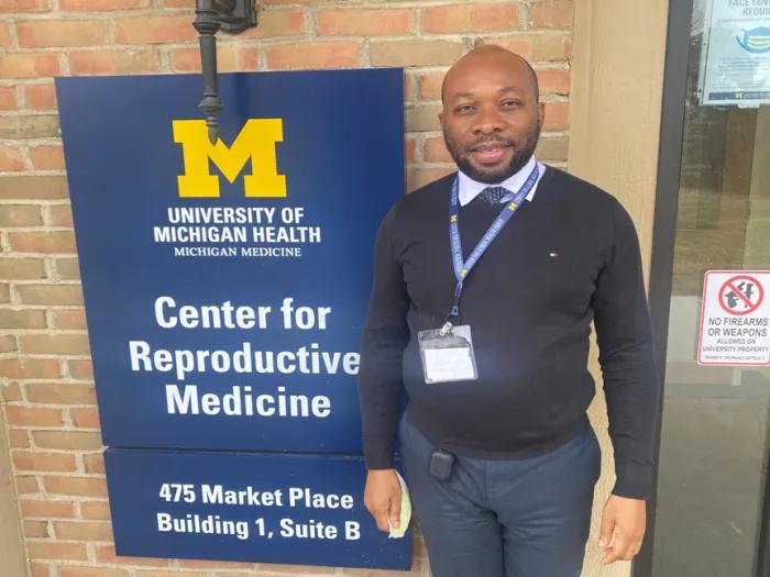 Man wearing a black sweater over a collared shirt, standing next to a sign for the Center for Reproductive Medicine