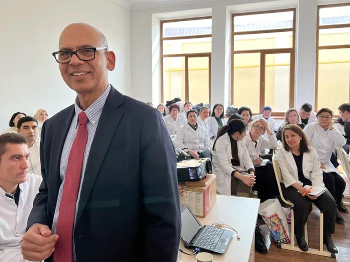 Man wearing glasses, blazer, and tie, standing in a room full of doctors in white lab coats
