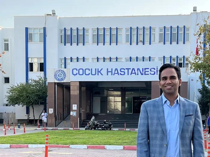 Man wearing a grey checked blazer standing outside a white building with Turkish words written across the front