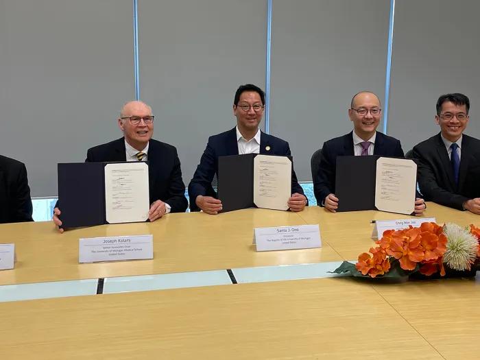 Five men in dark blazers and ties seated at a large conference table, with the three men in the middle holding up black folders with a paper in them