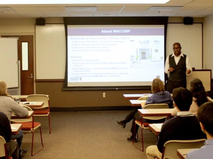 A man giving a presentation in the front of a classroom to other people sitting at desks