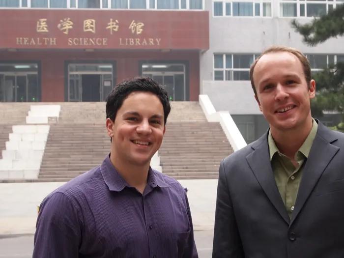 Two men in business clothes standing in front of a building with a maroon sign that reads 'Health Sciences Library' in English and Mandarin