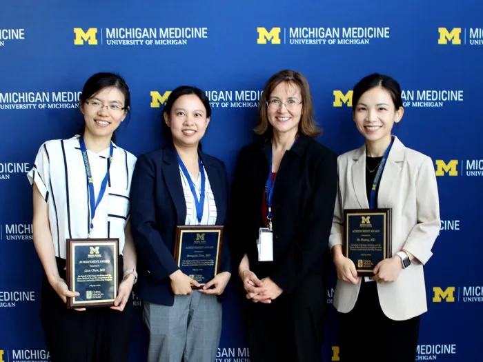 Four women standing in front of a blue wall that reads "Michigan Medicine," holding plaques