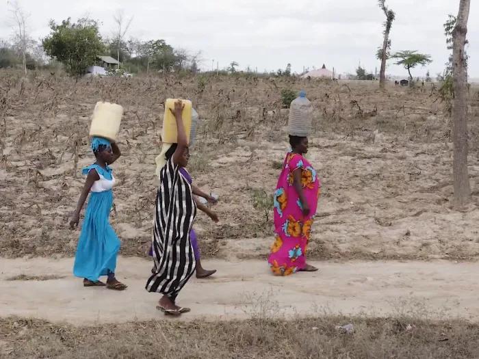 Four women in bright, patterned dresses, walking on a dirt road and carrying buckets on their head