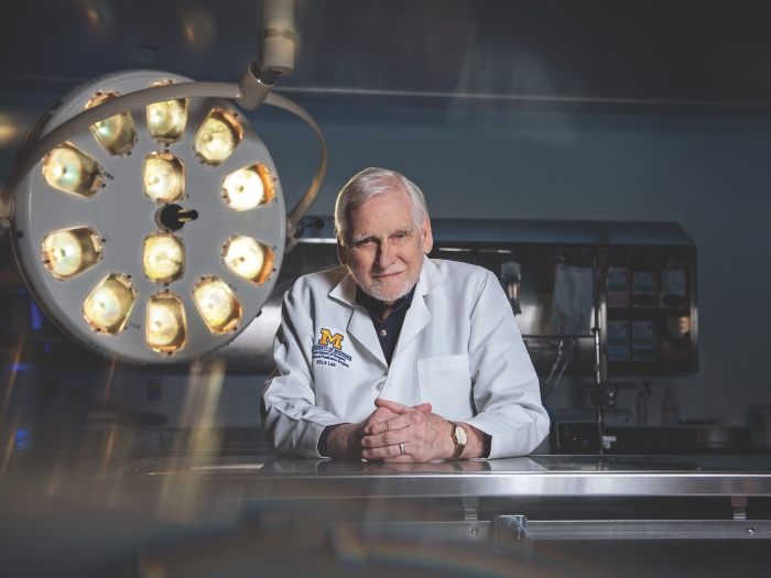 Dr. Robert Bartlett is wearing a doctor's white coat and leaning on a steel table in a lab with his hands clasped. He's standing next to a large circular surgical light. 