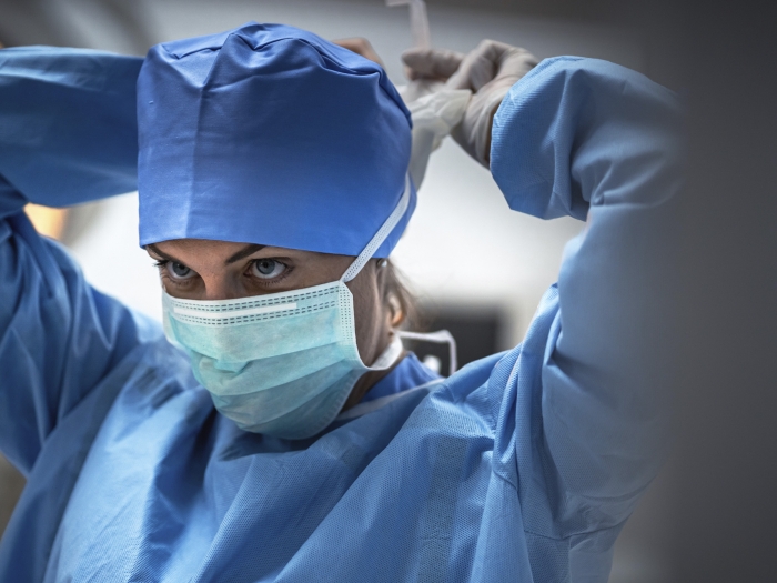 female surgeon in blue cap scrubs and mask tying mask and looking up 