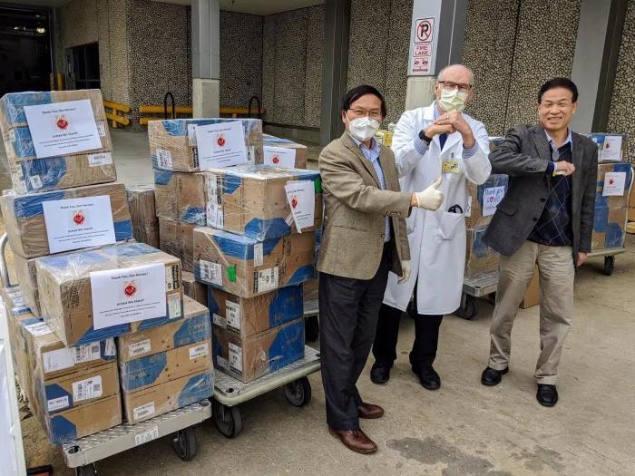 A man in a white lab coat wearing a face mask, a man in a brown blazer wearing a face mask, and another man in a black blazer all standing together in front of stacks of cardboard boxes 