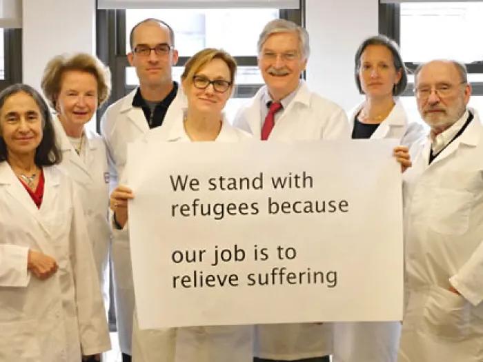 A group of people in white lab coats standing with a white sign that reads 'We stand with refugees because our job is to relieve suffering."