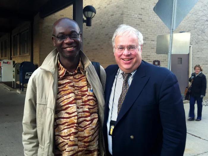 A man in a black blazer and tie standing next another man in a khaki jacket and tiger-striped-patterned shirt
