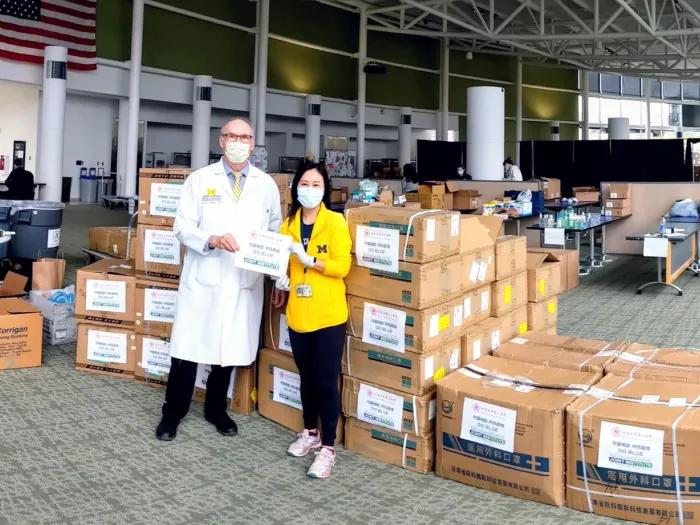 A man in a white lab coat and wearing a face mask standing next to a woman in a yellow jacket and also wearing a facemask. There are a lot of cardboard boxes stacked behind them