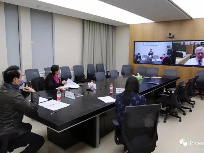 People sitting around a long table in a conference room, looking at a screen which is displaying a video call