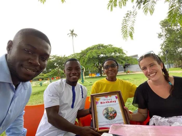 Four people sitting outside in a park, holding a framed 'Happy Birthday' card