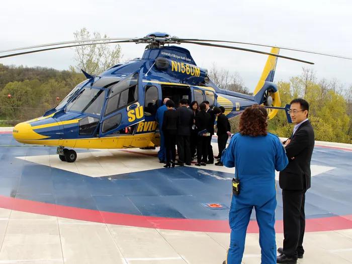 Maize and blue helicopter on a helipad, with a group of people standing on the helipad