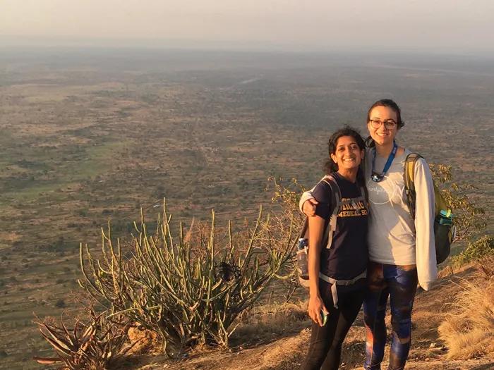Two women in athletic clothes and wearing backpacks standing on top of a sandy hill, with a more arid landscape in the background