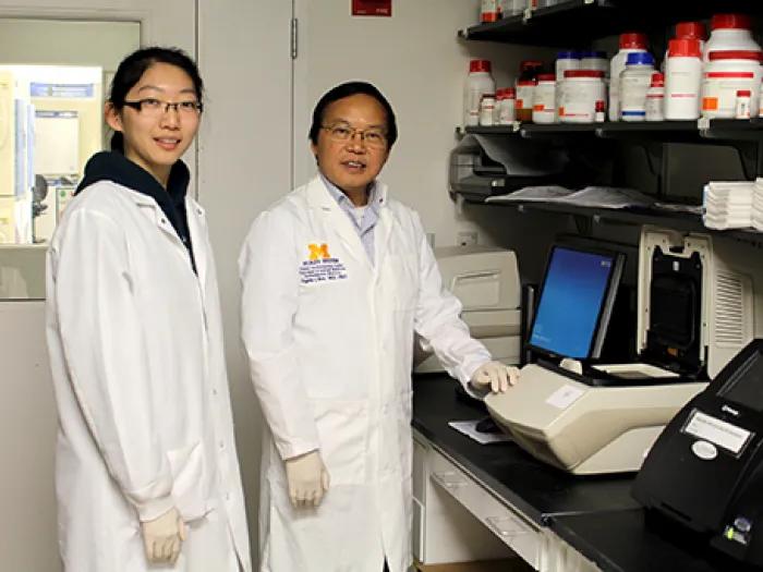 Two people in white lab coats, standing in a research lab next to some equipment 