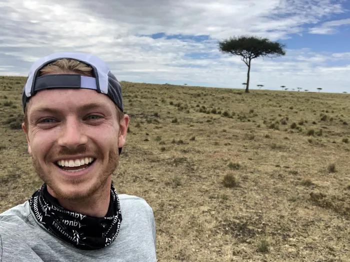 Man wearing a backwards baseball cap, grey shirt, and black cloth around his neck, with a savannah and tree in the background