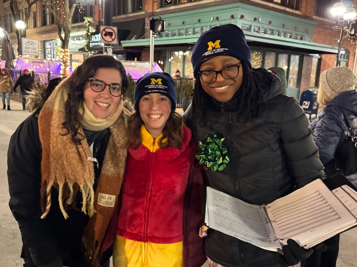 Three women wearing winter coats and holding sheet music stand outside in downtown Ann Arbor in the evening