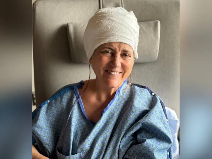 woman smiling after brain surgery with brain wrapped in white cloth and in blue hospital gown on grey background