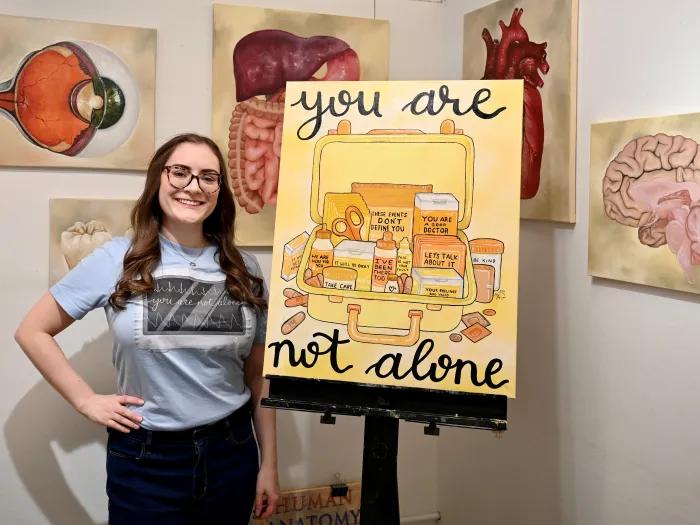 Courtney Burns standing next to the "You Are Not Alone" first aid kit painting by Morgan Graznow. 