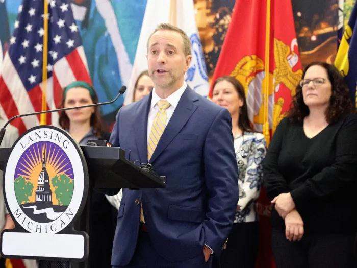 man speaking at podium with people standing behind him and flags