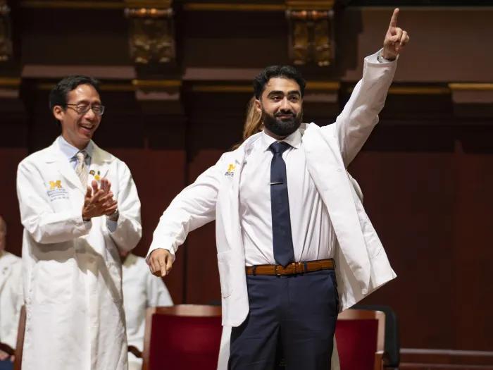 A medical student is cloaked in his white coat while pointing to his family and friends while medical school dean applauds