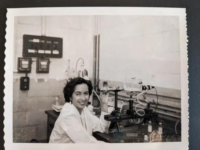 Black and white photo of Elaine Shafrin smiling and looking up from her work in a laboratory. A table full of lab equipment is in front of her.