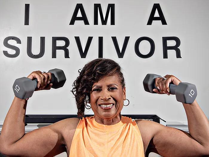 Black woman holds weights in front of I am a survivor sign